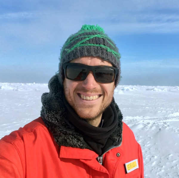 A man in a bright red waterproof jacket wearing a green beanie and sunglasses in Antarctica.