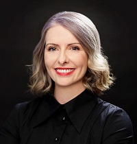 A portrait of a woman smiling at the camera. She is wearing a black top against a black background. She's wearing red lipstick and has silver hair.