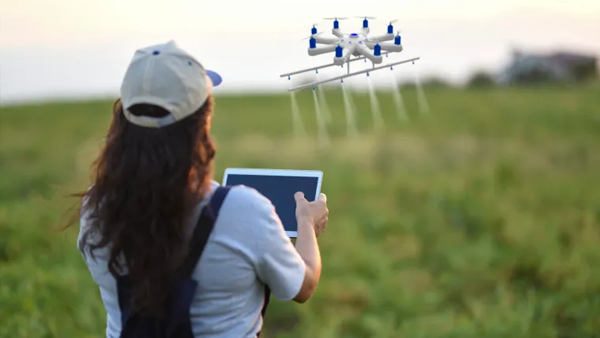 Woman wearing a baseball cap using a tablet to control a drone fitted with a spraying device