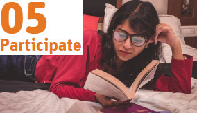 person with glasses lying on a bed reading a book