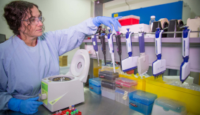 A female scientist in a white coat and safety goggles interacts with scientific equipment in a laboratory.