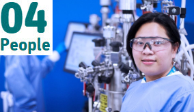 Woman in safety glasses standing against a machine smiling at the camera.