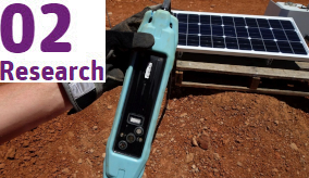 a hand holding a blue electronic device with a solar panel and red dirt in the background
